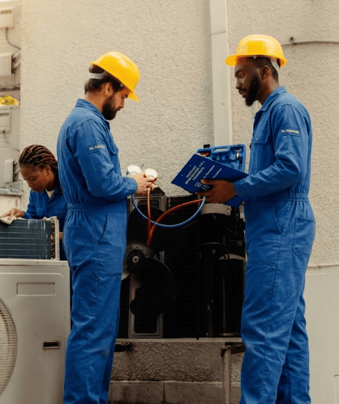 comparatif-primes-cvc-vs-frigoriste-idf Deux techniciens qualifiés en bleu de travail classique et casques jaunes effectuent une maintenance CVC/Froid sur une unité extérieure. L'un vérifie le manifold tandis que l'autre consulte une checklist lisible comparant les primes de salissure/itinérance (CVC) et de froid/astreinte (Frigoriste) en Île-de-France.