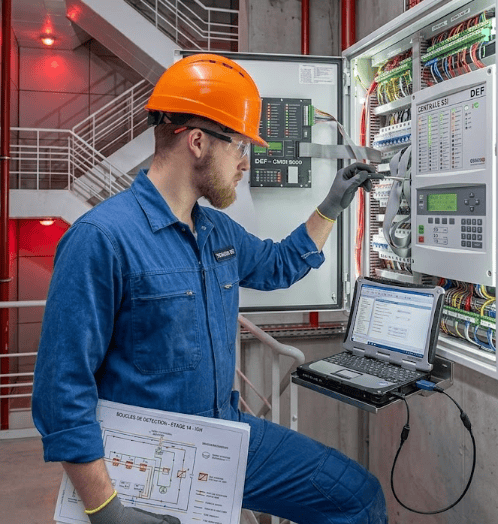 maintenance-ssi-expert-interim-idf-bref-service Technicien de maintenance spécialisé en sécurité incendie effectuant un diagnostic technique sur une centrale SSI DEF à l'aide d'un PC portable et d'un schéma de boucles de détection IGH en Île-de-France.